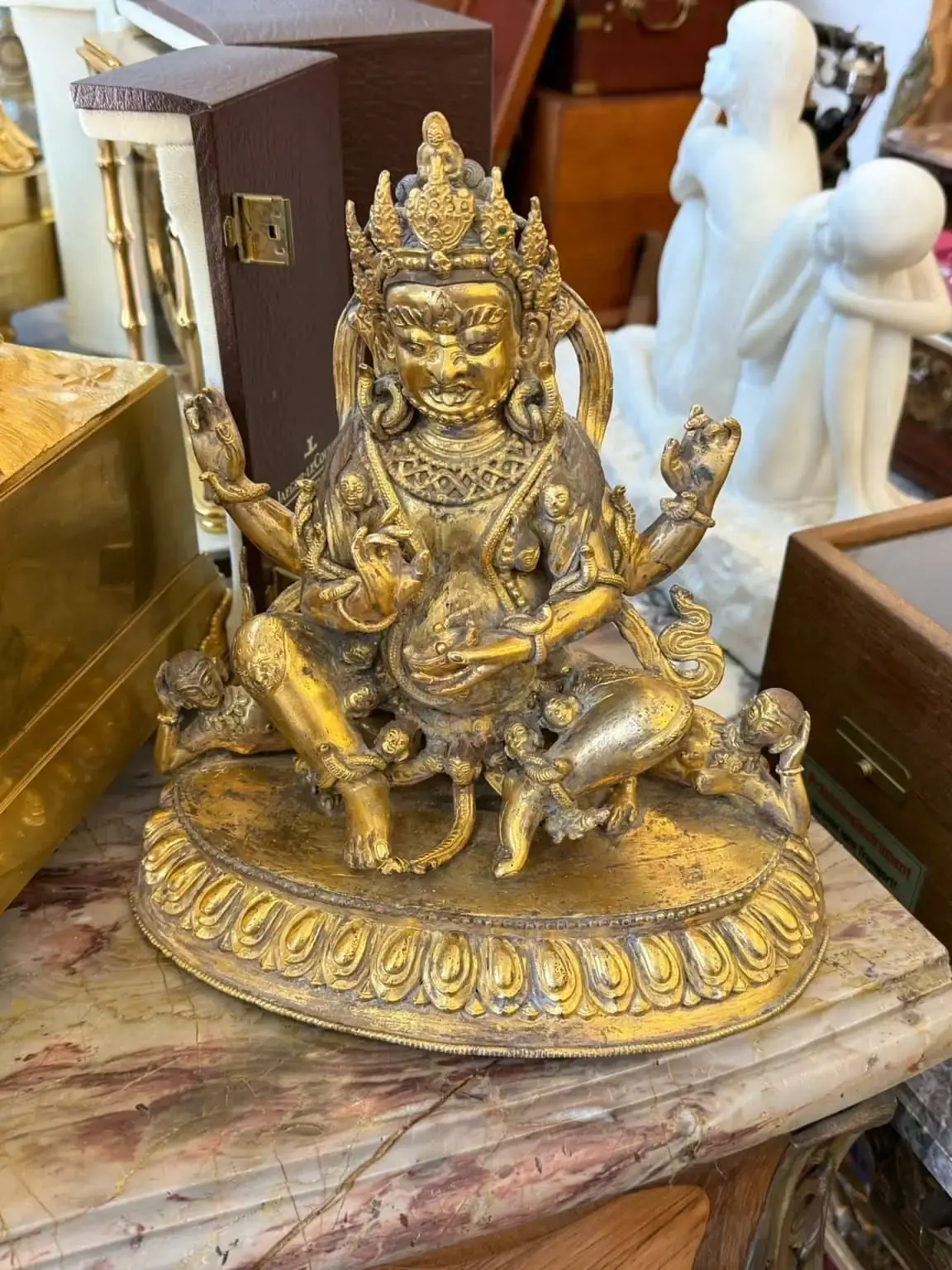 Une statue de divinité hindoue ou bouddhiste dorée détaillée avec plusieurs bras repose sur une base décorative, entourée d'autres objets vintage sur une surface en marbre dans un magasin d'antiquités à Lucerne.