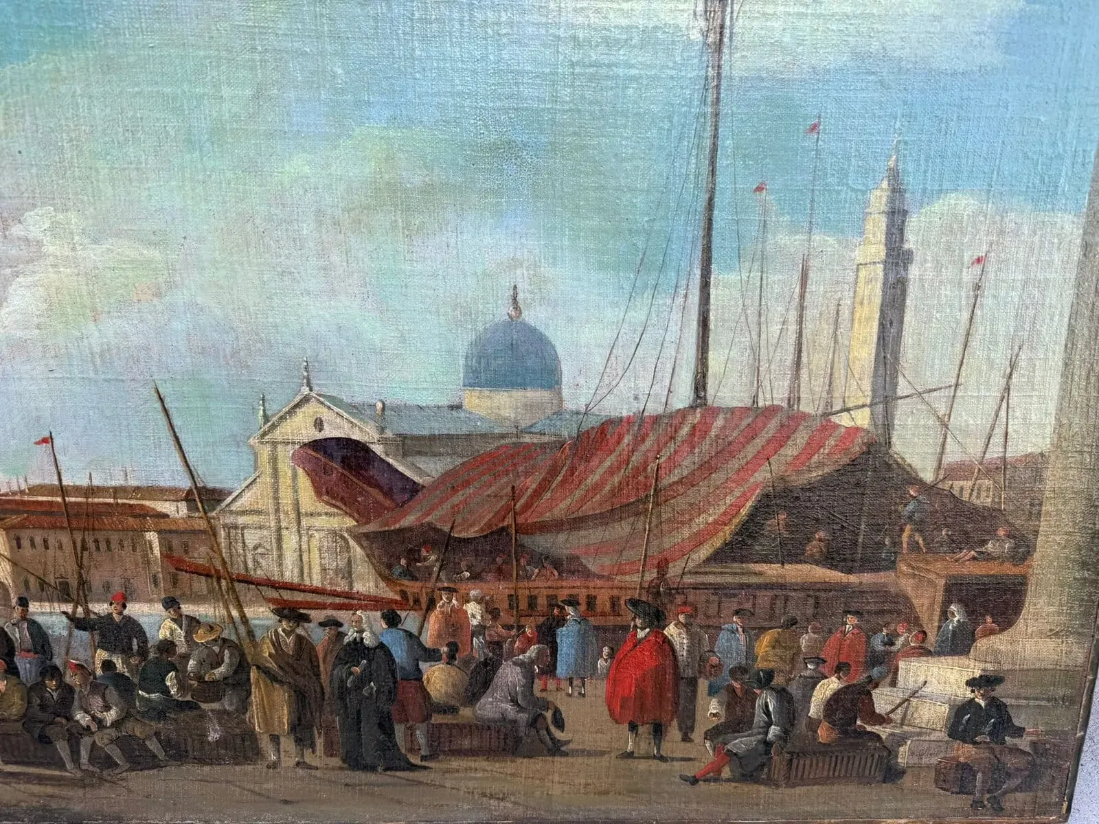 Une scène portuaire animée avec des gens rassemblés près des quais, des navires avec de hauts mâts, une tente rayée et des bâtiments historiques avec des dômes et des tours en arrière-plan sous un ciel partiellement nuageux.