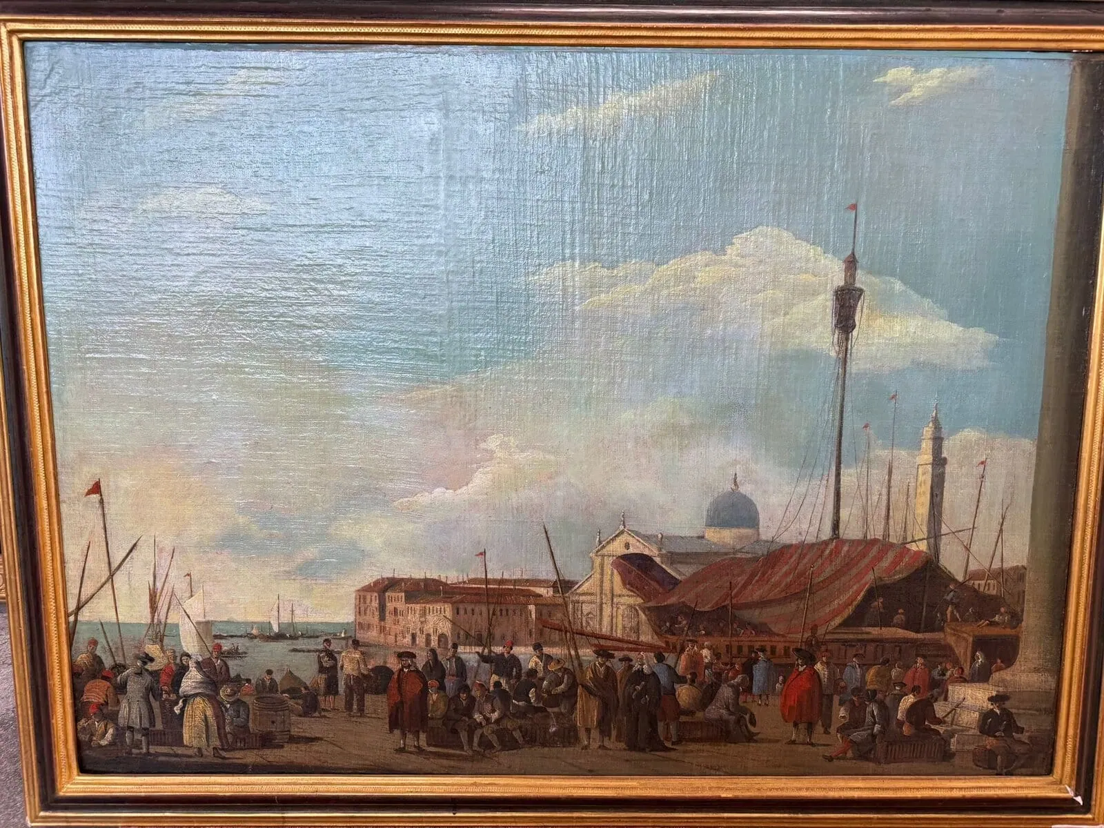 Tableau représentant une scène portuaire animée, avec foule, voiliers et tentes. À l'arrière-plan, des bâtiments historiques, des dômes et de hautes tours sous un ciel partiellement nuageux. Le cadre est doré.