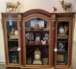 Une vitrine en bois et en verre présente diverses horloges anciennes ornées, dont une montre de Soleure, ainsi que des objets décoratifs. Au-dessus de la vitrine se trouvent deux figurines de vache et une de veau, tandis que d'autres horloges et objets décoratifs sont disposés à l'intérieur.