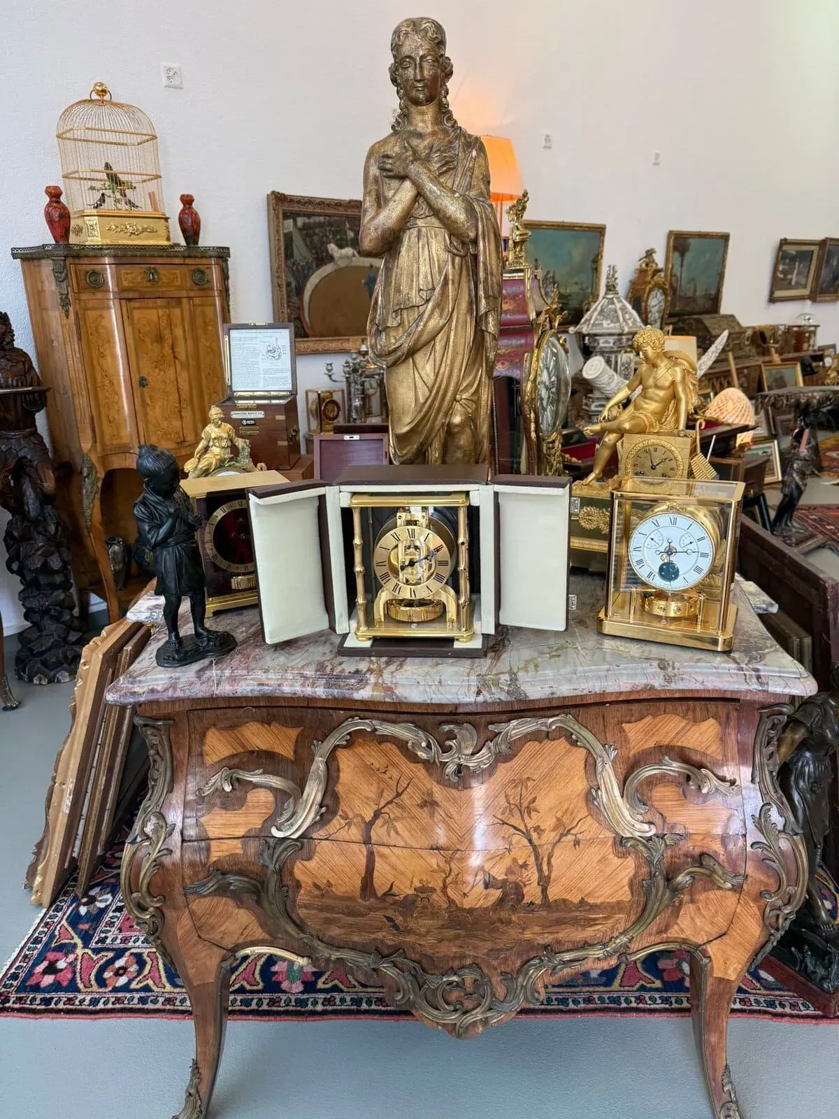 Un meuble en bois orné d'arbres sculptés contient des horloges anciennes et des statues en or. D'autres meubles anciens, des œuvres d'art encadrées et des objets décoratifs sont disposés derrière ce meuble dans un décor de magasin d'antiquités.