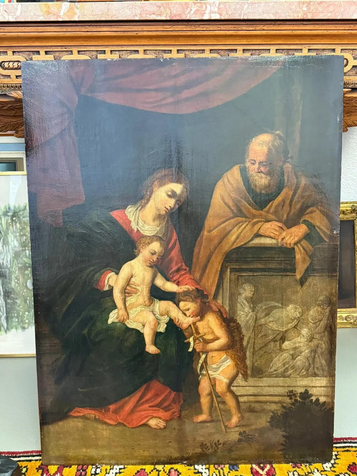 Une peinture classique représente la Vierge Marie avec l'enfant Jésus sur ses genoux, saint Joseph veillant sur eux, et un jeune Jean-Baptiste tenant une croix, sur un fond sombre et drapé et un relief de pierre.