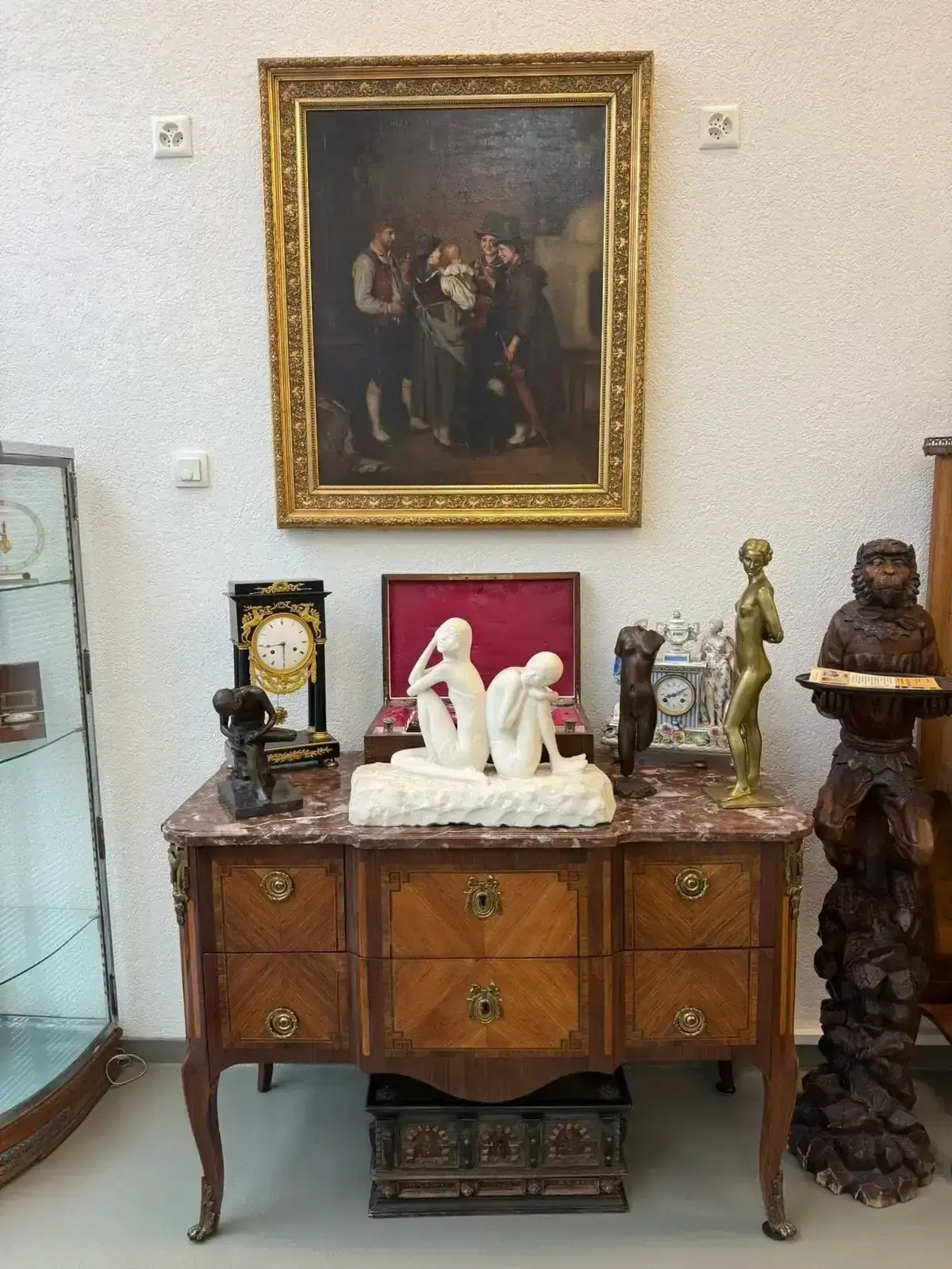 Un coffre en bois avec un plateau en marbre présente diverses sculptures, horloges et objets décoratifs. Il est surmonté d'un tableau encadré représentant des personnes en vêtements d'époque, évoquant l'élégance intemporelle que l'on retrouve souvent dans les maisons de Crans-Montana.