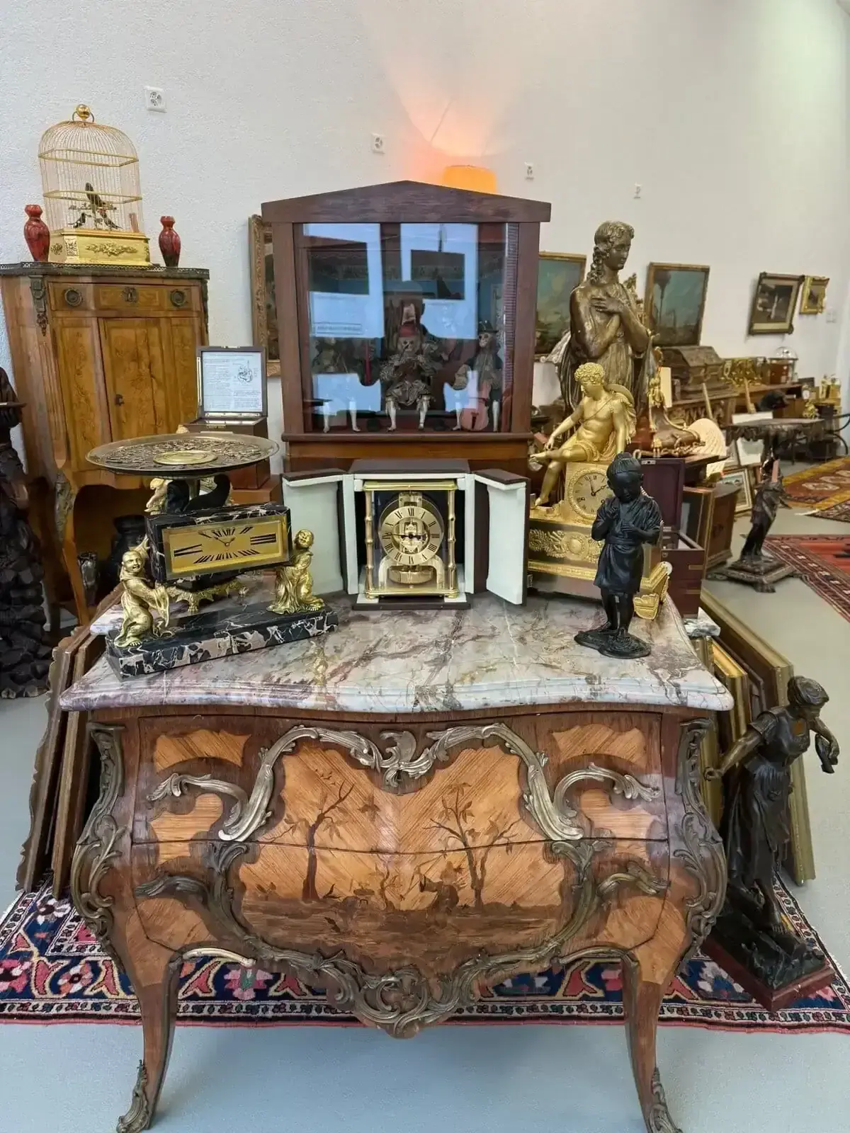 Dans une boutique de Crans-Montana, une antique commode en bois ornée de scènes peintes est posée sur des horloges, des statuettes et des objets décoratifs d'époque. D'autres antiquités, peintures et sculptures remplissent la pièce à l'arrière-plan.