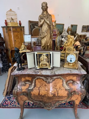 Un coffre antique en bois orné d'un motif d'arbre incrusté est surmonté d'horloges anciennes, d'une statue en or et de ce que j'achète - des trésors décoratifs. D'autres statues, objets d'art et lampes sont densément disposés, avec, en arrière-plan, une cage à oiseaux posée sur un meuble en bois.