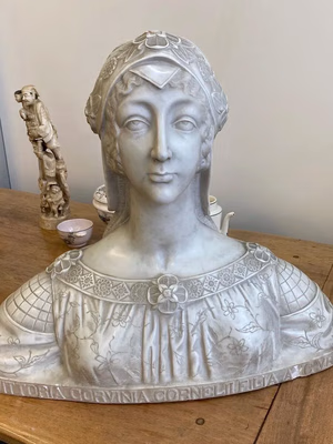 Buste en marbre d'une femme portant une coiffure complexe et une robe à motifs floraux et géométriques. Le buste est posé sur une table en bois, avec des tasses en porcelaine et une figurine à l'arrière-plan. Une inscription en latin est visible sur la base.