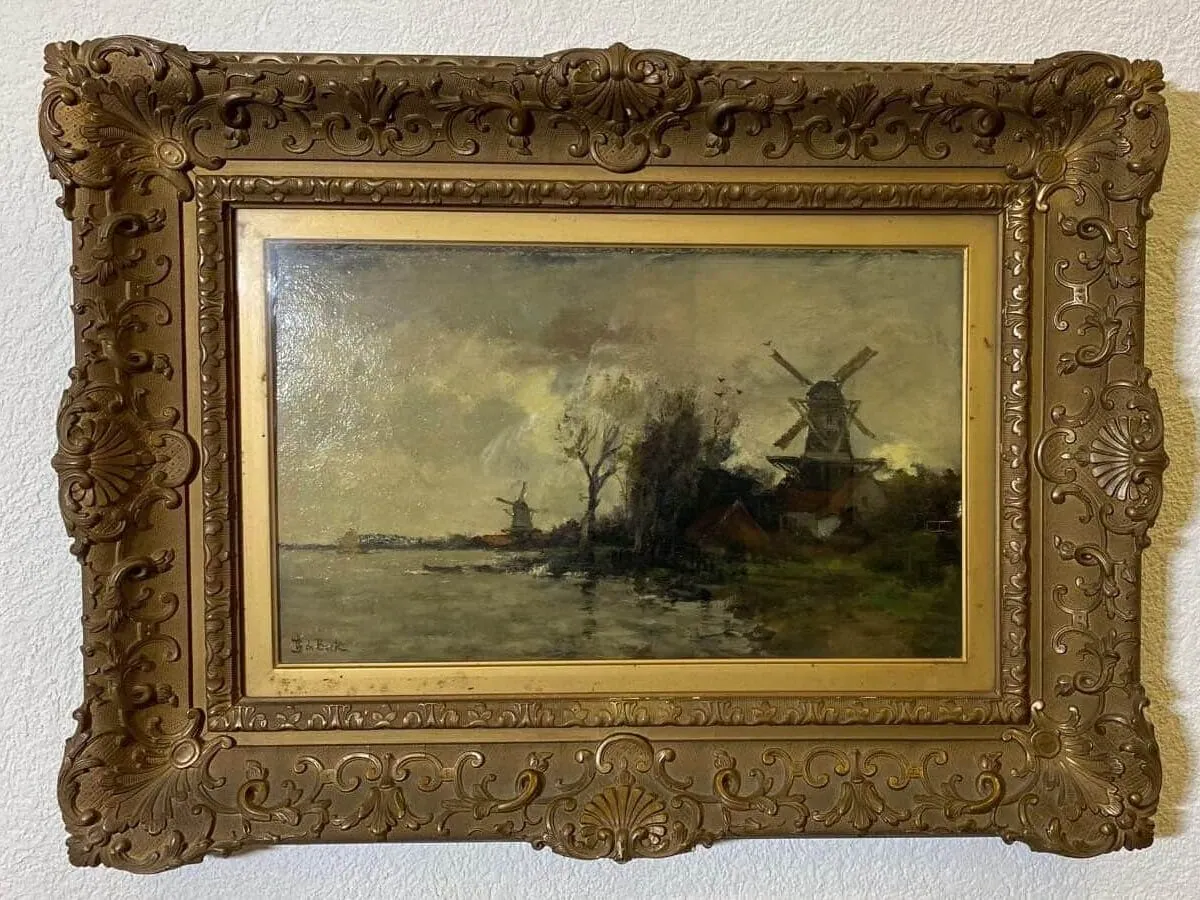 Un tableau encadré avec un cadre doré orné représente un paysage fluvial avec des arbres, des maisons et deux moulins à vent sous un ciel nuageux. L'eau reflète la scène et l'ambiance générale est sereine et légèrement sombre. Le tableau est accroché à un mur blanc.