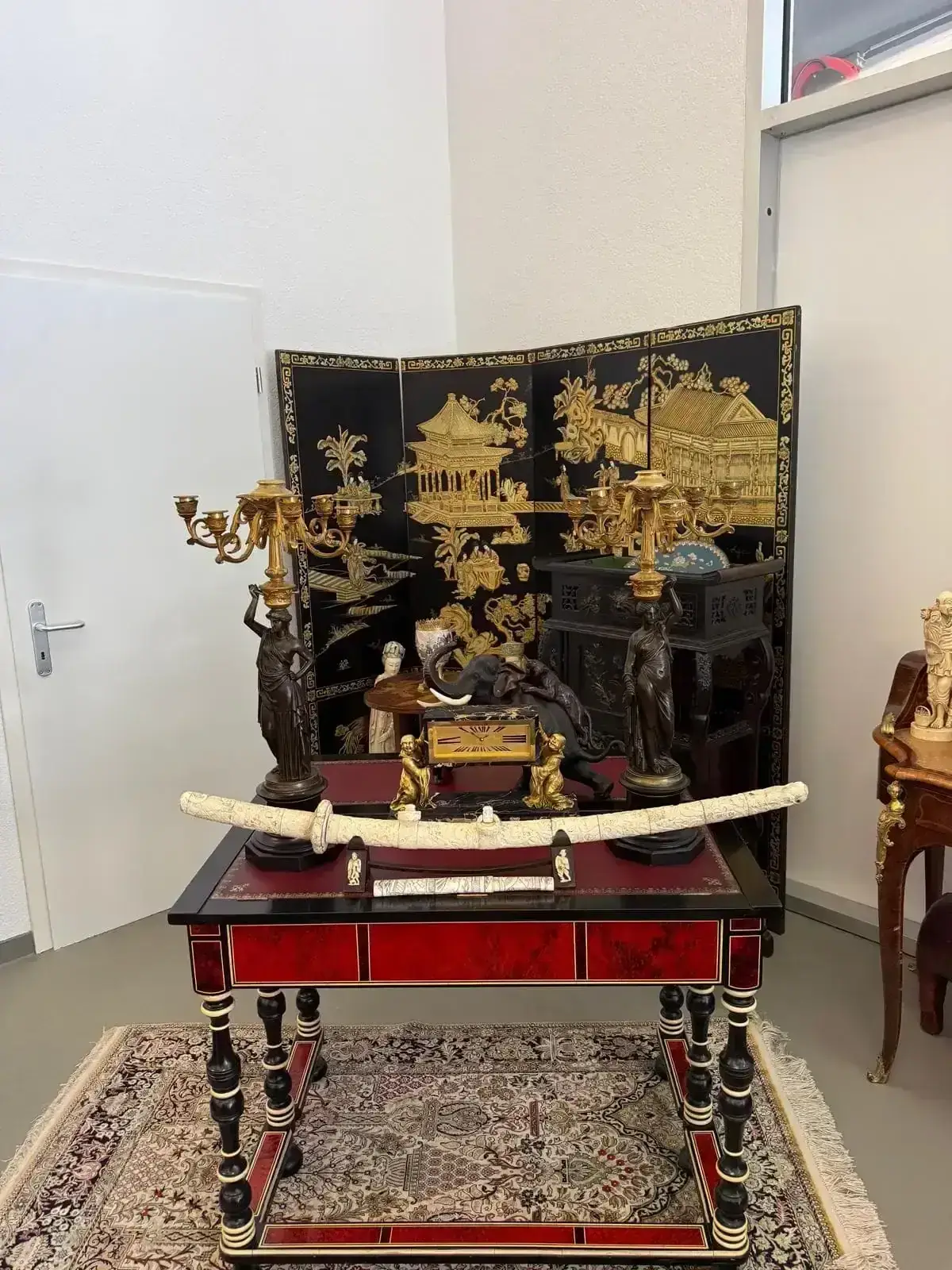 Une pièce décorative comprenant une table rouge et noire avec des accents dorés, surmontée d'une défense ornée, d'une horloge ancienne et de deux candélabres détaillés. Derrière, un paravent noir et or aux motifs asiatiques crée l'ambiance parfaite pour la dégustation de Bière.