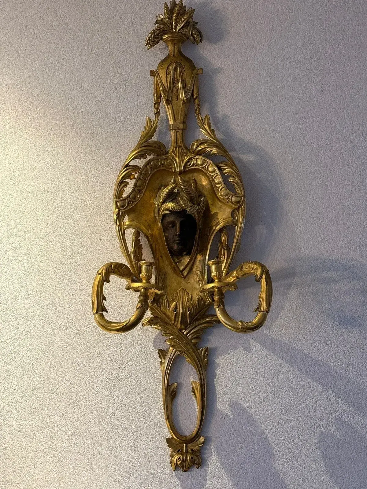 Une applique décorative Orbe ornée de motifs de feuilles complexes et d'un visage sculpté sombre, partiellement obscurci au centre, est suspendue à un mur blanc uni, projetant une ombre douce sur la surface texturée.