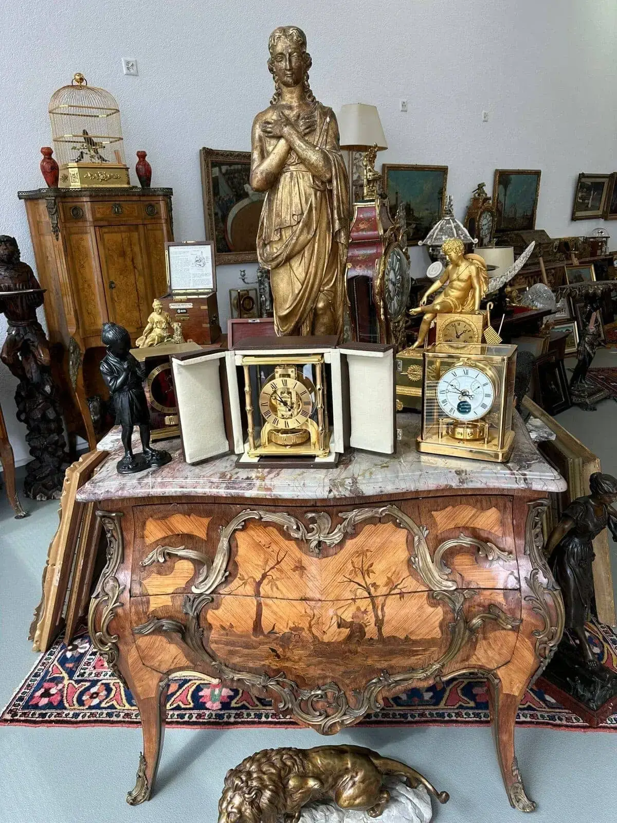 Une commode en bois ornée de sculptures complexes et d'une scène peinte supporte des statues en or, une horloge et une vitrine ouverte. À l'arrière-plan, des meubles anciens de La Neuveville, des œuvres d'art encadrées et une cage à oiseaux remplissent le magasin d'antiquités bondé.