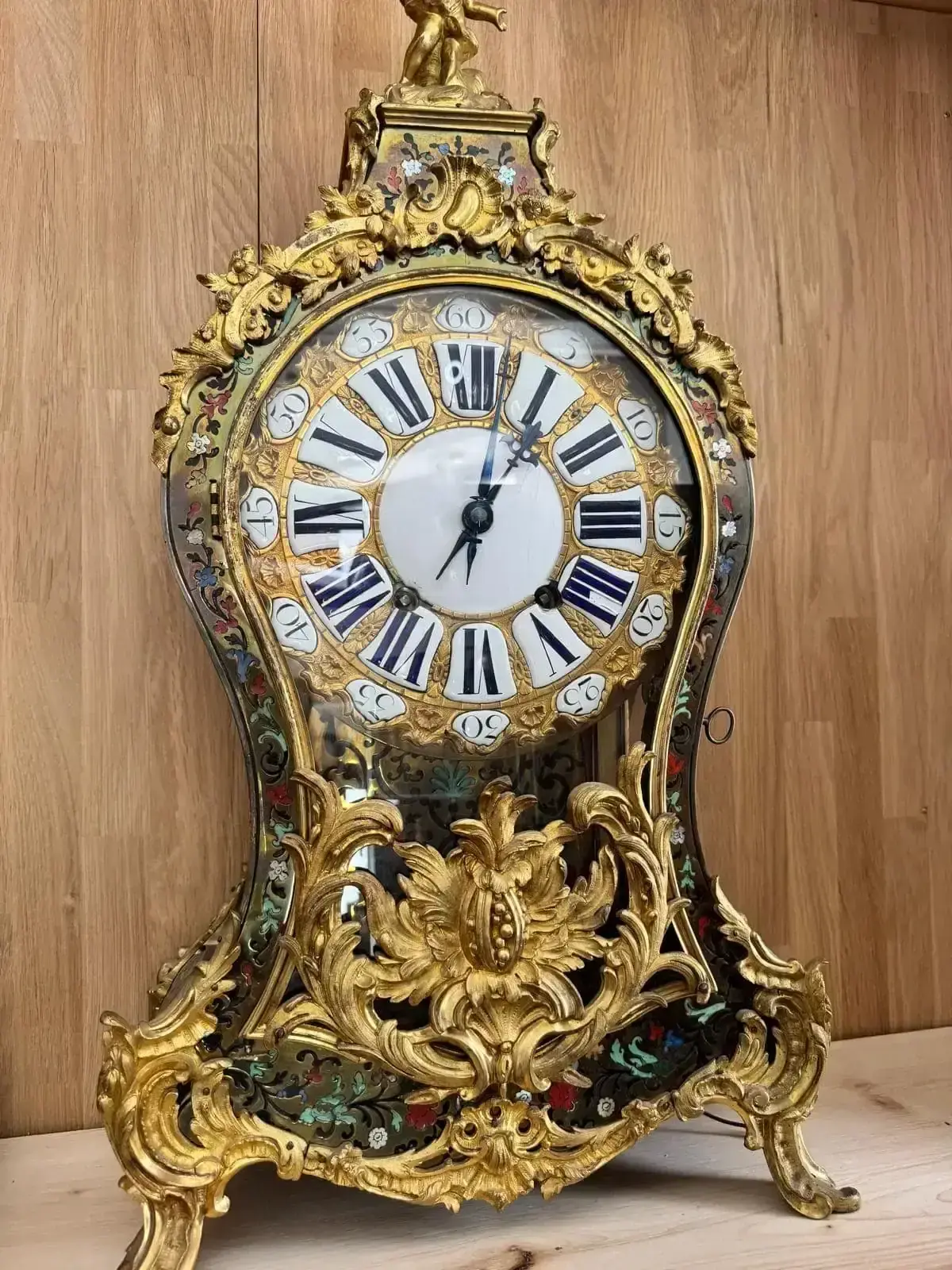 Une horloge ancienne ornée de détails dorés complexes et de motifs floraux repose sur une surface en bois clair, reflétant l'art des maîtres horlogers français et la riche histoire de l'horlogerie en France.