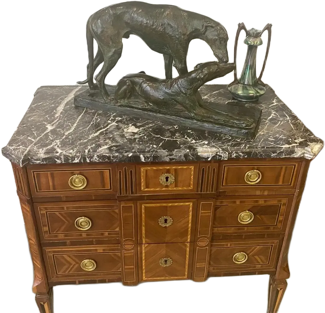 Une commode en bois avec un plateau en marbre foncé, avec des poignées en laiton et des incrustations décoratives dans le style d'Auvernier. Elle est surmontée d'une sculpture en bronze représentant deux lévriers et d'une petite cruche décorative en métal. La commode comporte plusieurs tiroirs avec des motifs en bois complexes.