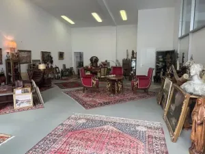 L'intérieur d'une boutique d'antiquités présente des meubles vintage, des chaises rouges tapissées autour d'une table, des tapis ornés, des œuvres d'art encadrées et des sculptures. Les murs blancs et l'éclairage lumineux créent une atmosphère spacieuse et accueillante.