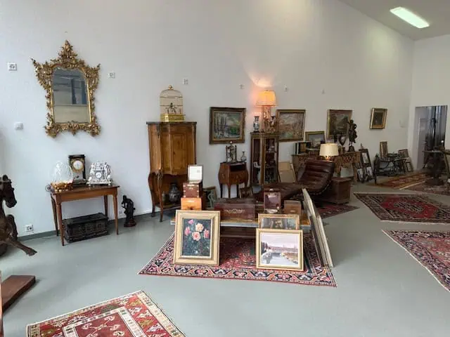 Une pièce avec des meubles anciens, dont un miroir décoratif, divers tableaux, une horloge et une lampe posée sur une armoire en bois. Plusieurs tapis sont disposés au sol et plusieurs œuvres d'art encadrées sont appuyées contre le mur du fond.