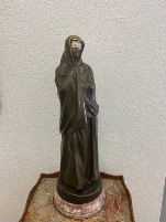 Une statue en bronze représentant un personnage encapuchonné se dresse sur un piédestal rond au socle en marbre, rappelant une pièce intemporelle que l'on pourrait acquérir pour ajouter de l'élégance à n'importe quelle pièce. Le mur uni et texturé en arrière-plan offre un contraste parfait.