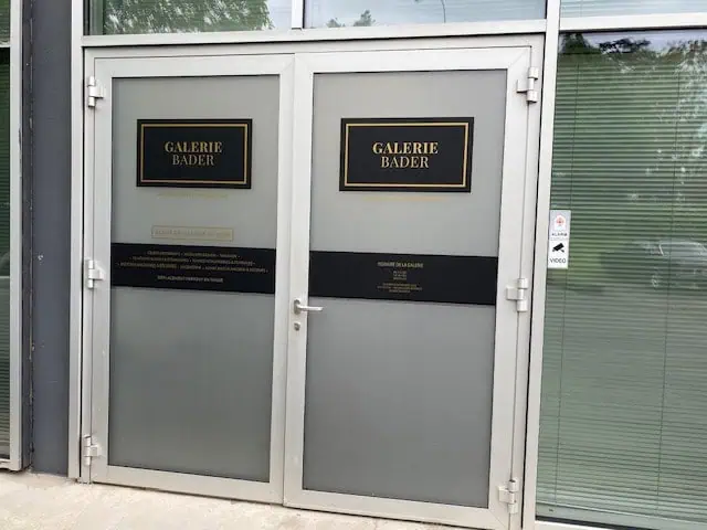 Une porte en verre grise ornée de deux panneaux noirs et dorés indiquant « Galerie Bader ». L'entrée fait partie d'un bâtiment moderne doté de grandes fenêtres. La poignée de porte est à gauche, et la fenêtre de droite est équipée de stores.
