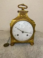Une horloge de cheminée vintage en or, avec un cadran blanc, des chiffres noirs et des motifs ornementaux, repose sur un tissu gris. Elle est ornée d'une poignée complexe et de petits pieds en forme de pattes de lion. Achetez cette exquise pièce d'horlogerie avant qu'elle ne disparaisse, avec la clé de remontage posée élégamment à côté.