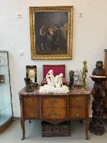 Une commode ancienne en bois avec un plateau en marbre présente diverses sculptures et horloges ; au-dessus est accroché un tableau encadré représentant trois personnes en vêtements à l'ancienne, évoquant l'élégance intemporelle des intérieurs historiques de Lucerne.