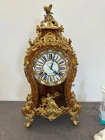 Une horloge ancienne ornée de détails décoratifs dorés, de chiffres romains et de sculptures complexes repose sur une surface à motifs. Inspirée du savoir-faire de La Chaux-de-Fonds, elle est ornée d'une petite statue sur son sommet et d'un vaporisateur visible à droite.