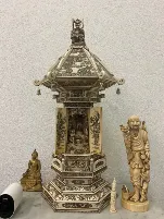 Un sanctuaire de couleur ivoire, inspiré de Soleure, finement sculpté, avec des motifs détaillés, une petite statue de Bouddha à l'intérieur, flanquée d'une figure en bois souriante et d'un Bouddha en or, le tout placé sur une surface en marbre contre un mur blanc texturé.