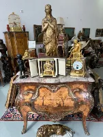 Un coffre antique en bois orné de sculptures et de peintures détaillées se trouve dans une pièce remplie d'objets anciens, notamment des statues en or, des horloges, des œuvres d'art encadrées, une cage à oiseaux et d'autres objets décoratifs.