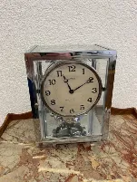 Une horloge Atmos vintage avec un cadre argenté et des côtés en verre est posée sur une surface en marbre. Provenant du canton de Zoug, son mécanisme est visible à travers le verre, tandis que le cadran de l'horloge indique 2:18 sur un mur blanc texturé.