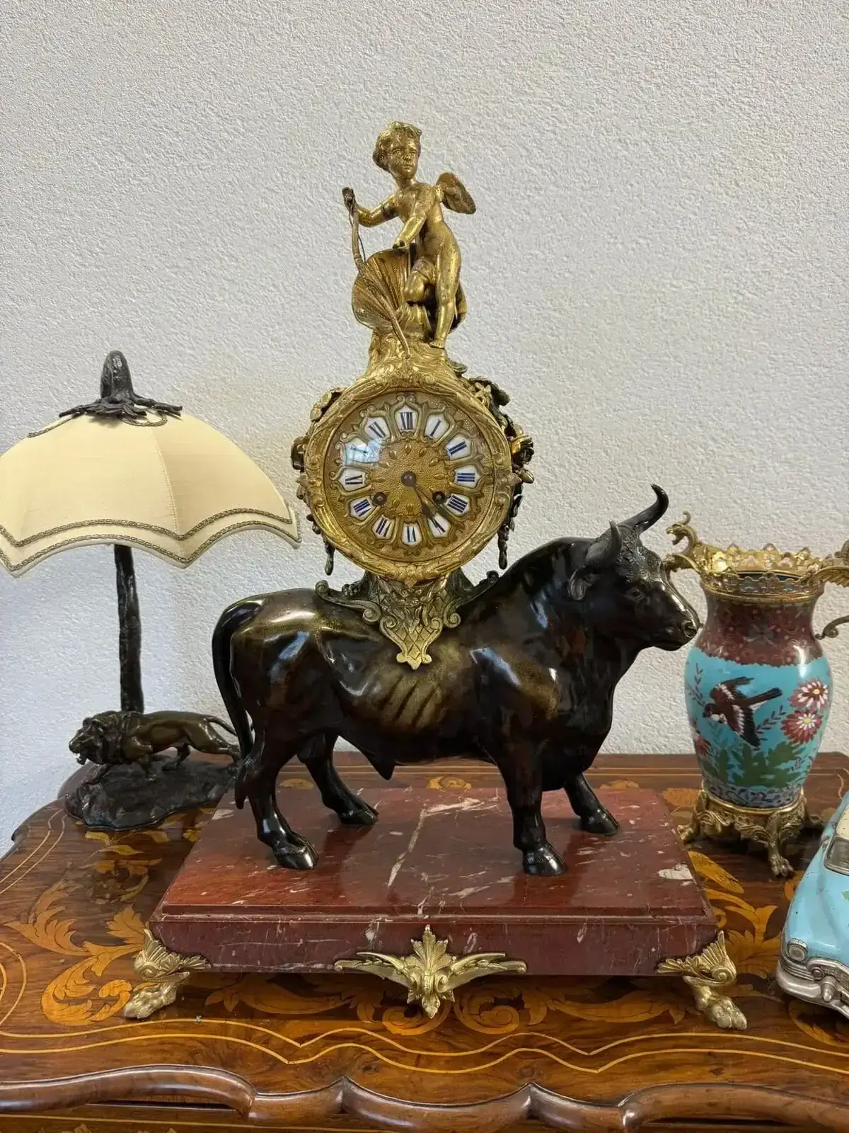 Une horloge de Versoix ornée de chiffres romains est montée sur une figurine de taureau en bronze. L'horloge est surmontée d'un chérubin en or avec une lyre. La base en marbre rouge repose sur une table en bois, entourée d'une lampe vintage, d'un vase et d'autres objets décoratifs.