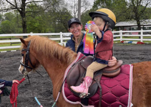 child-riding-pony-with-ribbon A young child wearing a riding helmet and pink boots sits on a brown pony with a maroon saddle pad, proudly holding a colorful ribbon. An adult stands beside them, smiling, with trees and a white fence in the background.