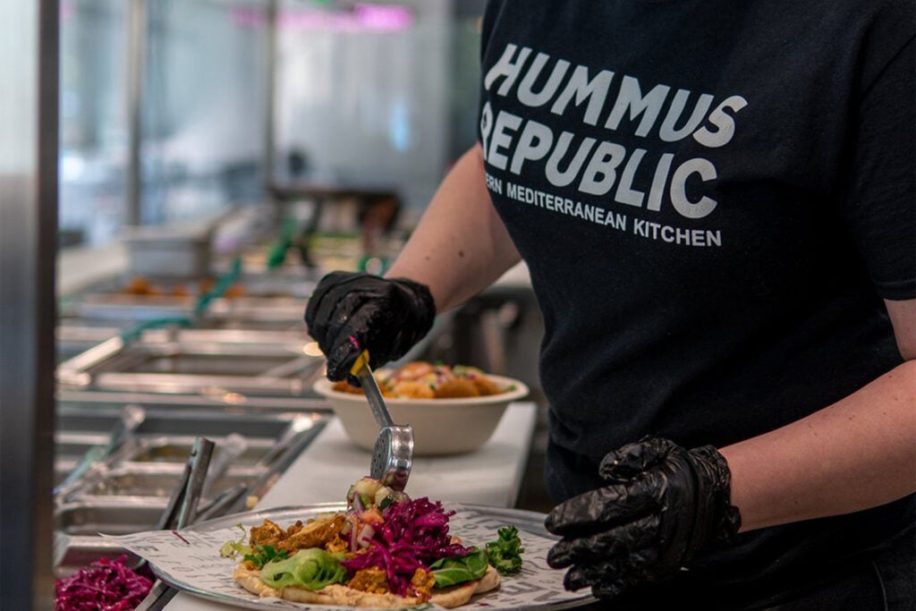 What Makes a Food Brand Feel Like Yours Before You Even Unlock the Door a person wearing gloves and a hummus republic shirt prepares a mediterranean dish at a food counter, adding toppings to a plate.