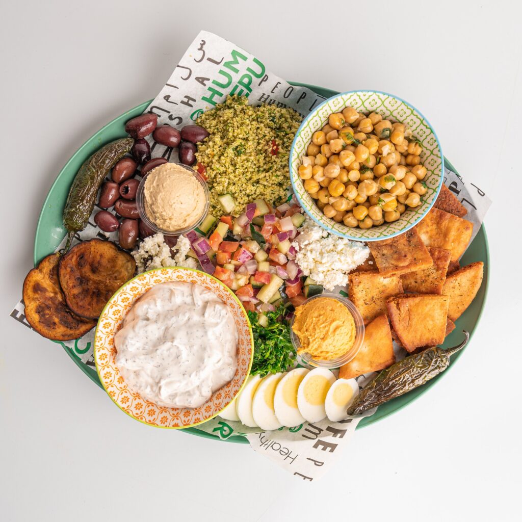 What It Means That the Food You Are Selling Is Food You Actually Grew Up Eating a platter with bowls of chickpeas, tabbouleh, tzatziki, and hummus, surrounded by pita chips, sliced eggs, plantains, olives, feta cheese, salad, and a grilled pepper.