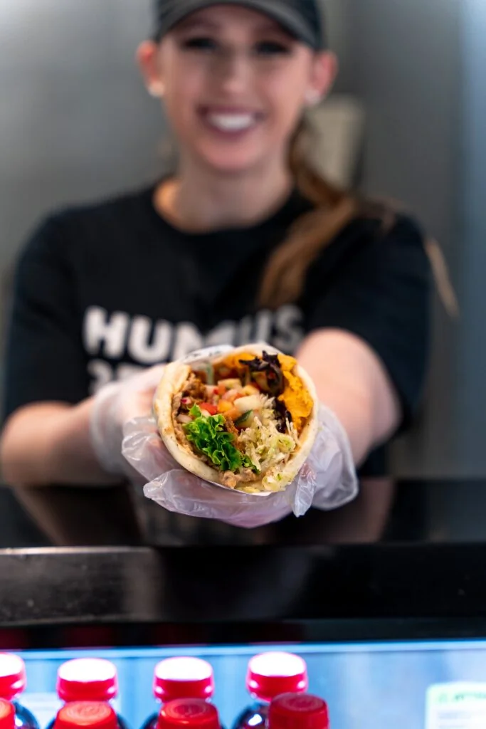 What Does It Really Cost to Open a Fast Casual Restaurant? An Honest Breakdown a person in a black shirt and cap, wearing gloves, holds out a filled pita wrap towards the camera; bottles are visible in a fridge below.