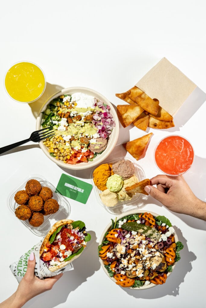 What Makes a Food Brand Feel Like Yours Before You Even Unlock the Door a variety of mediterranean dishes including a salad bowl, falafel, pita chips with dips, a wrap, two drinks, and a hand reaching for food on a white surface.