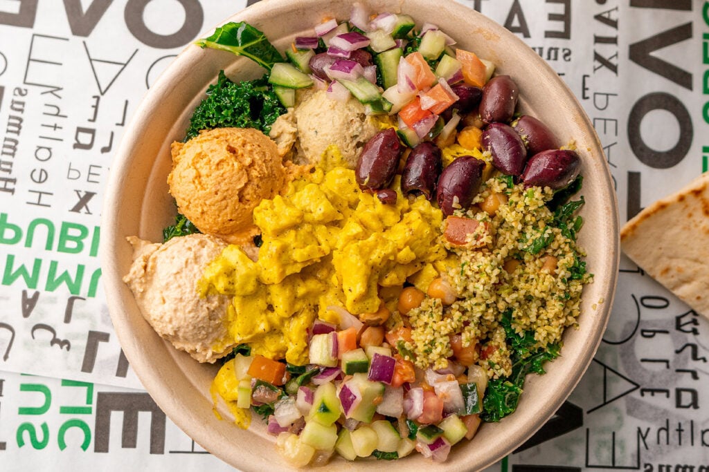 What a Manager-Run Location Model Actually Does to Your Income and Your Schedule a bowl with various mediterranean ingredients including hummus, tabbouleh, olives, chopped vegetables, greens, and a yellow sauce, on a graphic printed surface.