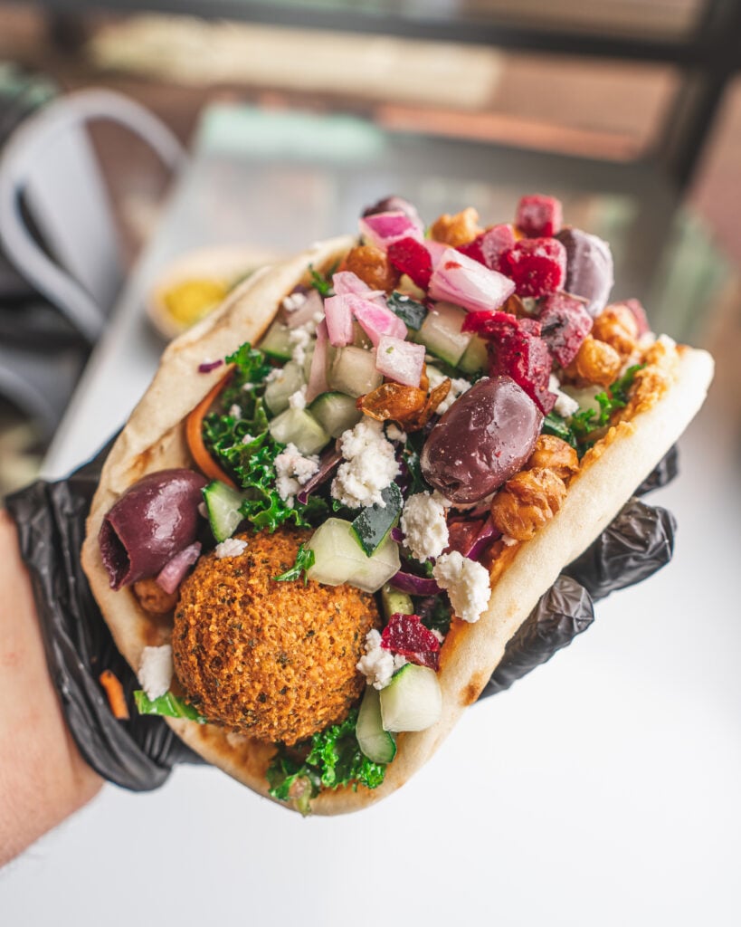 How a Centralized Supply Chain Actually Changes the Daily Reality of Running a Kitchen a gloved hand holds a pita wrap filled with falafel, cucumber, kale, olives, diced beets, feta cheese, and pickled onions.