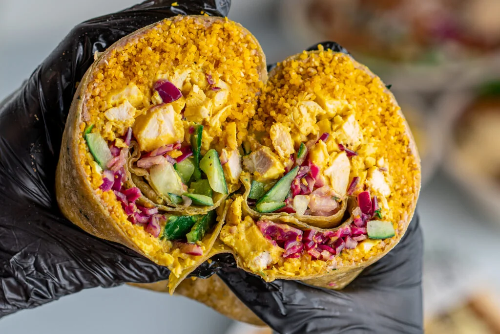 How First-Generation Owners Use Their Restaurant to Stay Connected to Their Roots a gloved hand holds a wrap cut in half, showing yellow rice, diced chicken, red onions, cucumbers, and lettuce inside.