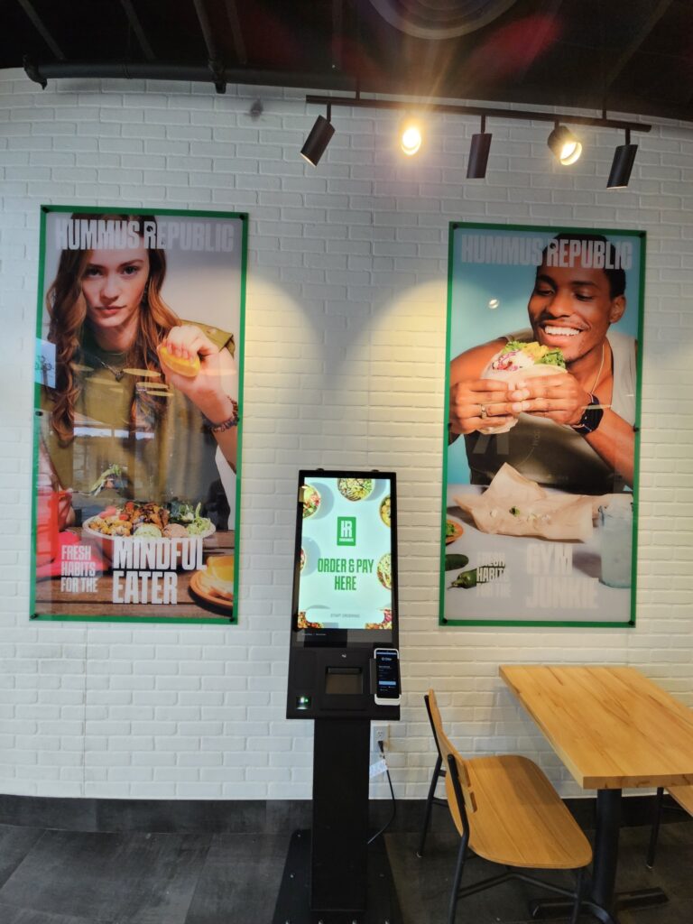What Does It Really Cost to Open a Fast Casual Restaurant? An Honest Breakdown two large posters of people eating food are displayed on a white brick wall above a self service kiosk and a wooden dining table in a restaurant.
