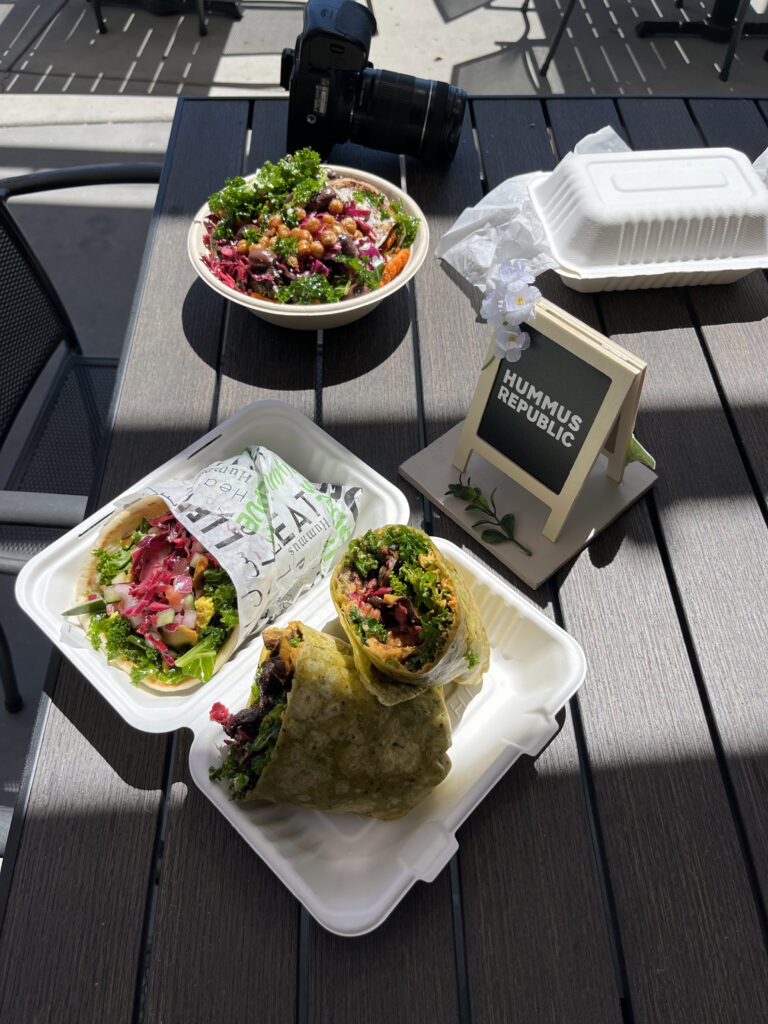 What Does It Really Cost to Open a Fast Casual Restaurant? An Honest Breakdown two takeout wraps and a salad bowl sit on an outdoor table with a camera, a takeout container, and a small hummus republic sign nearby.