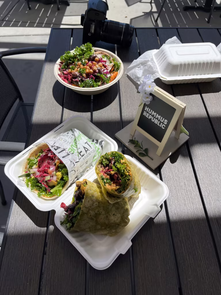 What Does It Really Cost to Open a Fast Casual Restaurant? An Honest Breakdown two takeout containers with wraps and a salad bowl are on an outdoor table, along with a camera and a small sign reading hummus republic.