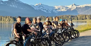 Explore Alaska on Two Wheels: The Ultimate iRide Alaska e-Bike Tour 1 Group with e-bikes posing by an Alaskan lake with snow-capped mountains in the background.