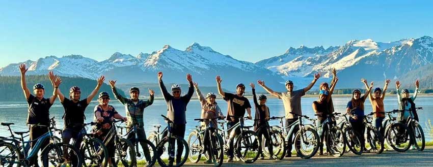 Explore Alaska on Two Wheels: The Ultimate iRide Alaska e-Bike Tour 4 Group with e-bikes raising arms beside an Alaskan lake with snow-capped mountains in the background.