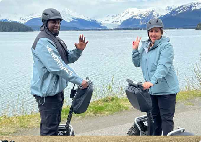SEGWAY Two people on Segways waving by the water with snowy mountains in the background during an iRide Alaska tour.