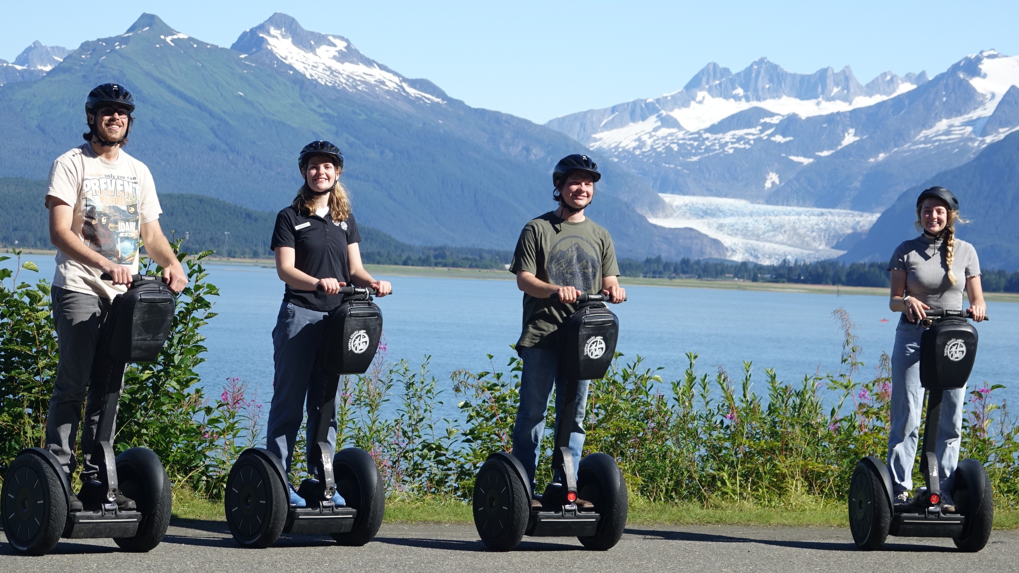 16.9 Two people on Segways waving by the water with snowy mountains in the background during an iRide Alaska tour.