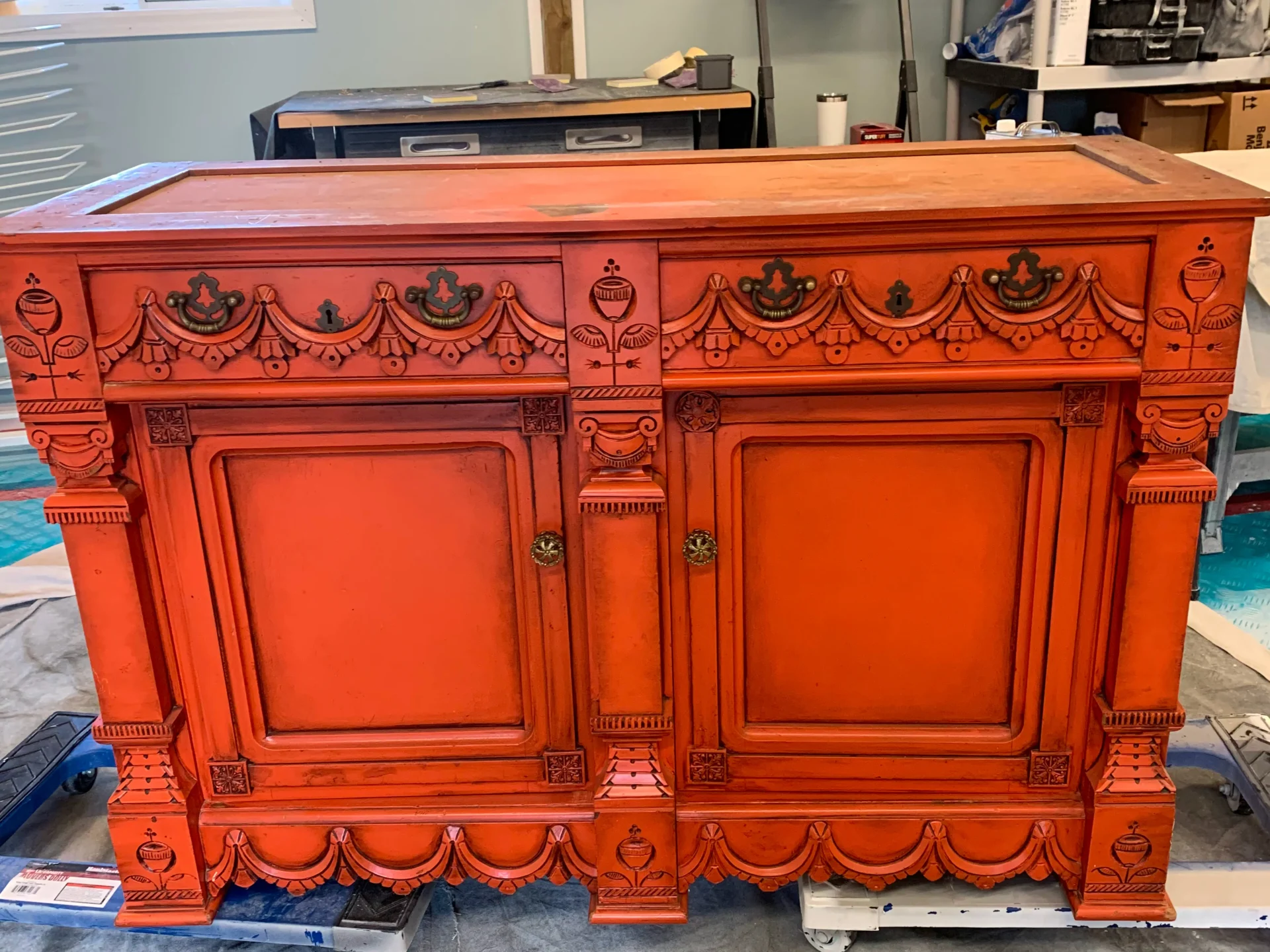 An ornate wooden cabinet painted bright orange with decorative carvings, two doors, and gold knobs, set in a workshop environment. - Jeff Bryant Painting