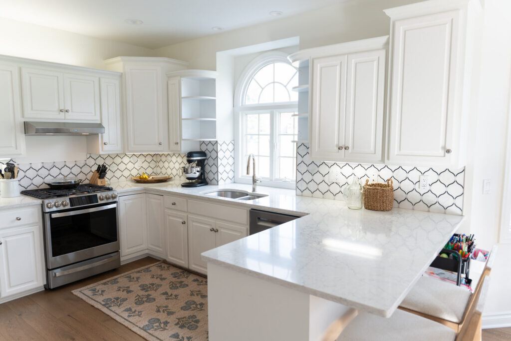 Modern kitchen with white cabinets, stainless steel appliances, a marble countertop, geometric tile backsplash, and fresh interior painting by Alto Michigan professionals. A window above the sink fills the space with natural light. - Jeff Bryant Painting