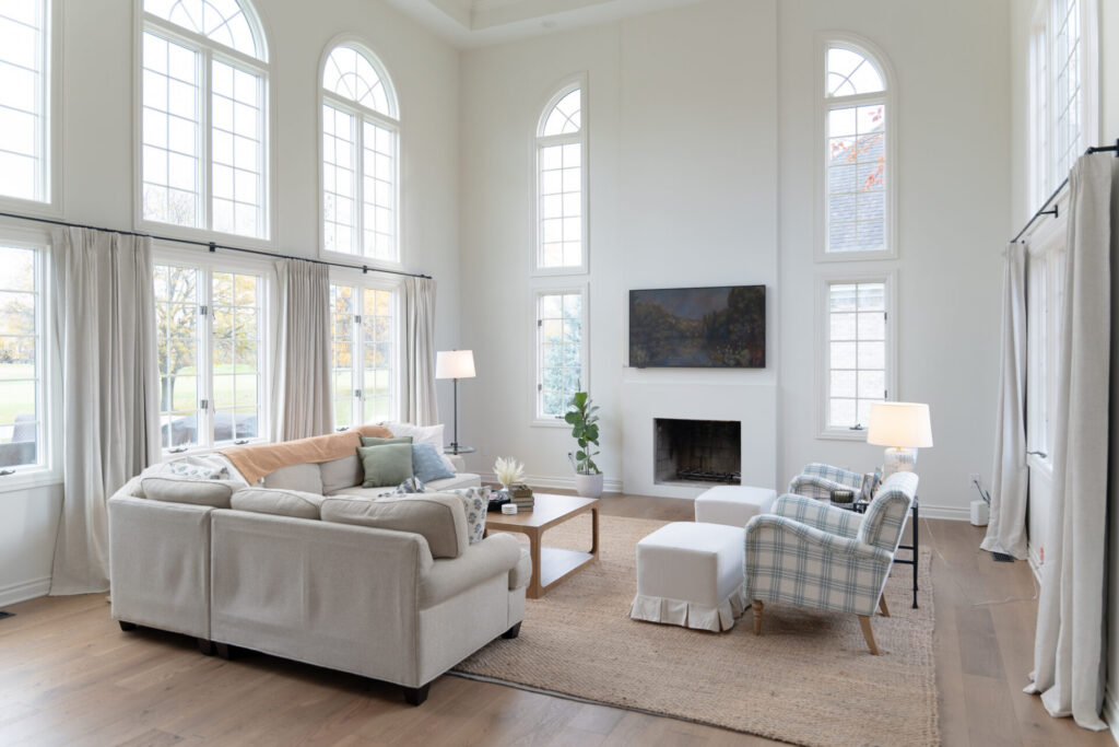 Spacious living room with tall arched windows, neutral sofas, a checkered armchair, a fireplace, and fresh interior painting—perfect for cozy nights in Alto, Michigan. Woven rug on wooden floors completes the inviting look. - Jeff Bryant Painting