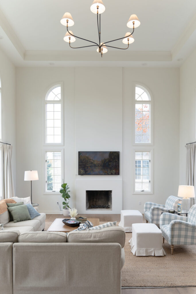 Bright living room with tall windows, a beige sectional sofa, two plaid armchairs, and a fireplace. Fresh interior painting in Alto Michigan enhances neutral tones, while natural light and a chandelier create an airy atmosphere. - Jeff Bryant Painting