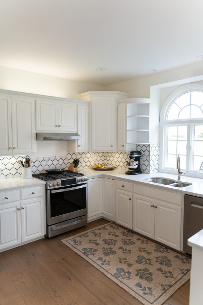 Bright kitchen with white cabinets, stainless steel appliances, patterned backsplash, a window above the sink, and a floral rug on light wood flooring—expert interior painting in Alto Michigan enhances every detail. - Jeff Bryant Painting