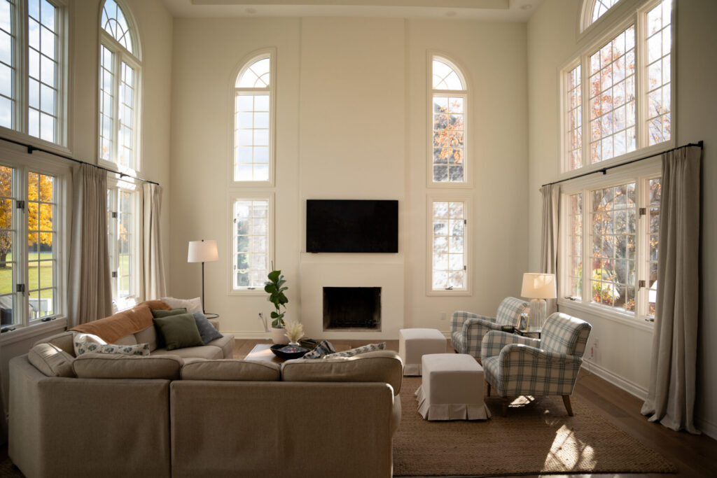 A spacious living room with high ceilings, large windows, a sectional sofa, two plaid armchairs, a fireplace, and a wall-mounted TV—beautifully refreshed by recent interior painting in Alto Michigan. Sunlight streams through the windows. - Jeff Bryant Painting
