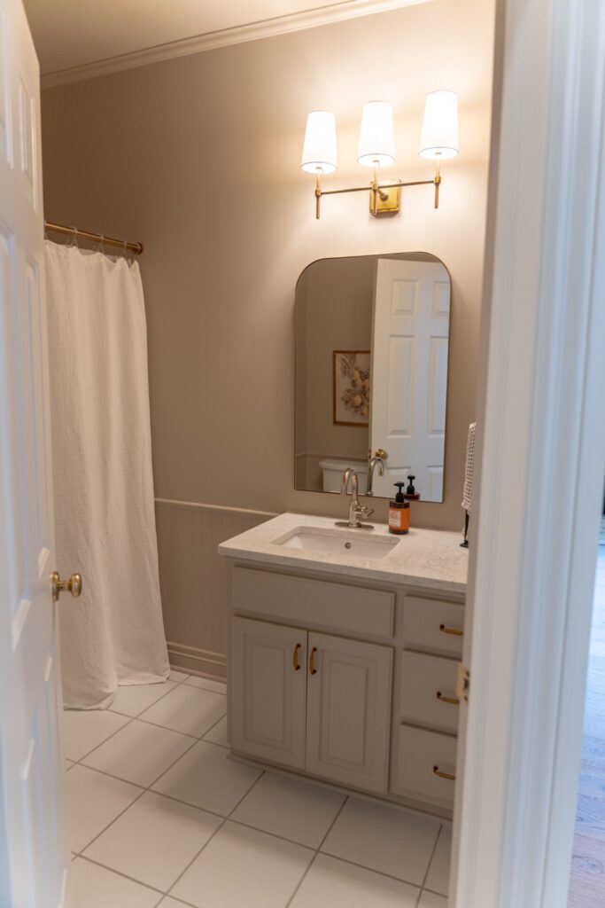 A bathroom with a white shower curtain, a vanity with a sink, a mirror, and two wall-mounted lights above the mirror. Fresh interior painting in Alto Michigan complements the bright white tile floor. - Jeff Bryant Painting
