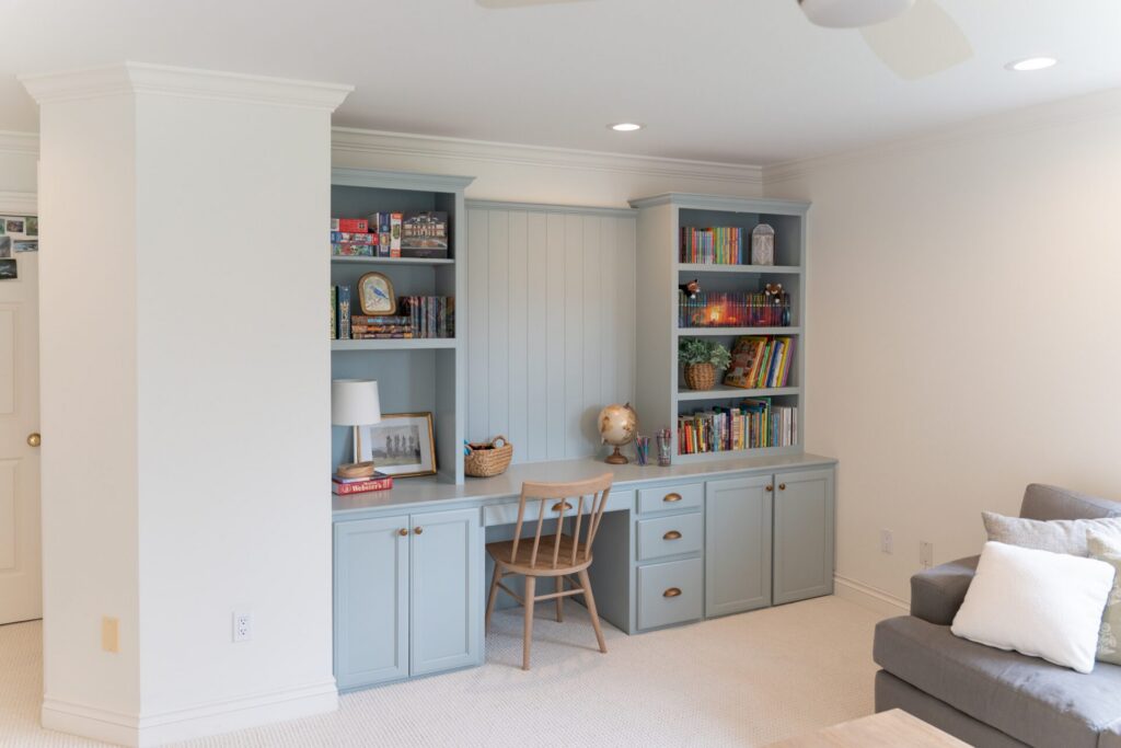 Built-in light blue shelves and desk, enhanced by expert interior painting Alto Michigan, hold books, games, a lamp, and a globe with a wooden chair beside a gray sofa in a bright room. - Jeff Bryant Painting